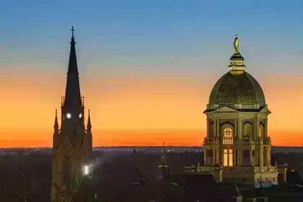 Sunset over the University of Notre Dame campus, with the Golden Dome and the Basilica of the Sacred Heart silhouetted against an orange and blue sky.